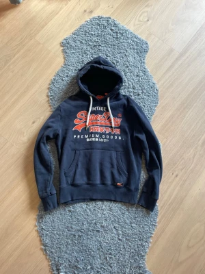 Mörkblå Superdry hoodie med tryck - Snygg mörkblå hoodie från Superdry med stor logga och japanskt tryck i vitt och orange på bröstet. Klassisk känguruficka framtill och vita dragsnören i huvan. Perfekt för en avslappnad streetwear-look.