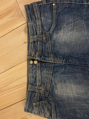 Blå jeans med dubbla knappar - Säljer ett par blå jeans med snygg slitning och dubbla knappar i midjan. Jeansen har kontrastsömmar, fem fickor och coola svarta broderier på bakfickorna. Klassisk passform och tillverkade i denim med en autentisk tvätt.