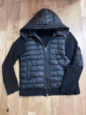 Svart cardigan från Moncler - Snygg svart cardigan från Moncler med quiltad kropp och stickade ärmar. Jackan har huva, dragkedja framtill och Moncler-logga på ärmen. Perfekt för kyliga dagar och ger en sportig men stilren look.
