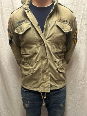 Field jacket Ralphlauren - Fieldjacket Ralph Lauren med patchar på ärmarna och fyra fickor framtill. Jackan har hög krage, axelklaffar och dragsko i midjan. Perfekt för dig som gillar utility-stil och detaljer.