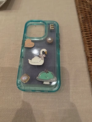 Turkost mobilskal med svan och dekorationer - Snyggt turkost mobilskal i genomskinlig plast med dekorativa motiv som svan, hjärta, pärlor och en tårtbricka. Skalet har en unik och lekfull design som sticker ut och passar dig som vill ha något personligt till mobilen. För iPhone 16 pro och bokstaven E går att ta bort!