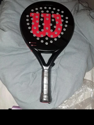 Wilson padelracket svart/röd - Säljer ett padelracket från Wilson i svart med stora röda W-loggan på träffytan. Racketen har rund form och många hål i bladet för bättre kontroll. Handtaget är lindat med svart grepptejp. Perfekt för dig som vill ta ditt padelspel till nästa nivå.