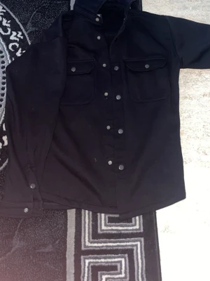 Svart overshirt med tryckknappar - Stilren svart overshirt med två bröstfickor och tryckknappar framtill. Overshirten har klassisk krage och raka ärmar med knapp vid ärmslut. Perfekt att bära över en t-shirt för en avslappnad look.