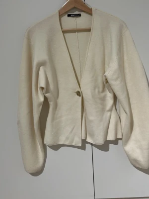 Krämvit kofta från Glamorous - Stilren krämvit kofta från Glamorous med v-ringning och en dekorativ knapp framtill. Koftan har långa ärmar och lätt utsvängd midja för en snygg siluett. Perfekt att slänga över en topp för en enkel men chic look.