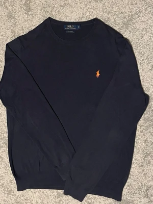 Mörkblå tröja från Polo Ralph Lauren - Snygg mörkblå tröja från Polo Ralph Lauren med klassisk orange broderad logga på bröstet. Tröjan har rund halsringning och långa ärmar. Perfekt för dig som gillar stilrena och tidlösa plagg.