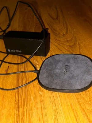 Mophie trådlös laddningsplatta - Säljer en svart trådlös laddningsplatta från Mophie med tillhörande strömadapter. Plattan har en mjuk yta i tyg och rundad fyrkantig form. Perfekt för att ladda din mobil utan sladdar. Enkel att använda och snygg design som passar på skrivbordet.