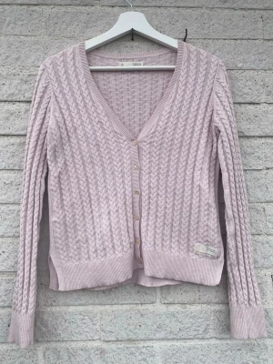 Ljusrosa kabelstickad kofta Odd molly - Söt kabelstickad kofta i ljusrosa från Odd molly. Koftan har v-ringning, ribbade muddar och knappar framtill.  Tröjan har en gullig rosett på ena ärmen och har andra fina detaljer. Perfekt att slänga över en topp för en chill look. Materialet känns mjukt och stretchigt. Knappast använd. I amerikansk storlek är det: 0 men skulle säga att den passar någon i storlek Xs/S