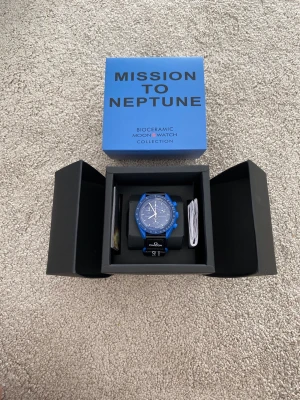 Omega x Swatch MoonSwatch Neptune - Säljer en blå Omega x Swatch Mission to Neptune från Bioceramic Moonwatch Collection. Klockan har en sportig design med blå boett och matchande blå urtavla, samt svart textilband med Omega-logga. Perfekt för dig som gillar unika och färgstarka accessoarer.