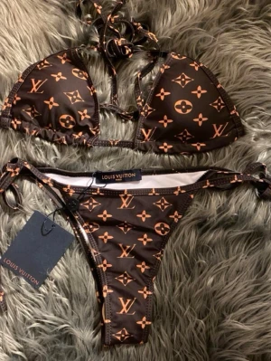 Brun bikini från Louis Vuitton - Snygg brun bikini från Louis Vuitton med klassiskt monogrammönster i beige. Bikinin har trekantsmodell på överdelen och knyt i både rygg och sidor. Perfekt för sommaren och strandhäng! Helt oanvänd, säljs då den inte passade