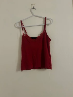 Röd linnetopp med spetskant - Snygg röd linnetopp med tunna axelband och dekorativ spetskant vid urringningen. Perfekt att styla med jeans eller kjol för en trendig look. Materialet känns mjukt och lätt, toppen har en enkel och rak passform.