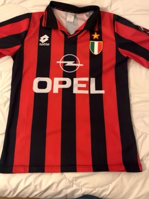 AC Milan retro matchtröja Lotto - Snygg AC Milan retro matchtröja från Lotto med klassiska röda och svarta vertikala ränder. Tröjan har Opel-logga på bröstet, klubbmärke med stjärna och italiensk flagga, samt nummer 3 och texten 'Palestine' på ryggen. Tillverkad i lätt och ventilerande syntetmaterial.