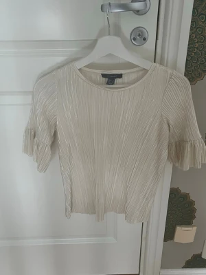 Plisserad ljusbeige topp från Primark - Säljer en plisserad ljusbeige topp från Primark med korta ärmar och volangdetaljer. Toppen har rund halsringning och ett glansigt material som ger en elegant känsla. Perfekt att styla med jeans eller kjol för en trendig look.