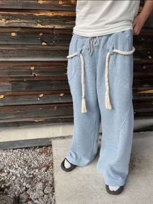 Ljusblå mjukisbyxor med rep - Unika ljusblå mjukisbyxor med bred passform och grova rep som dragsko i midjan och vid fickorna. Byxorna har stora fickor och är gjorda i mjukt sweatshirtmaterial, perfekta för en chill och avslappnad stil.