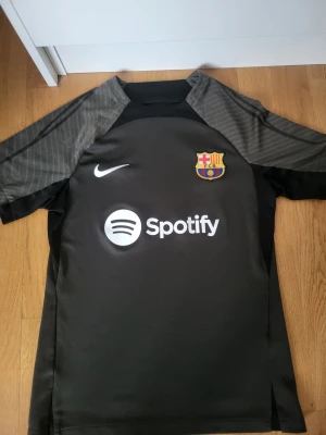 Svart FC Barcelona Nike träningströja - Snygg svart FC Barcelona träningströja från Nike med Dri-FIT-material. Tröjan har klubbmärke, Nike-logga och Spotify som sponsor framtill. Slim fit-modell med grå detaljer på ärmarna. Perfekt för fotbollsträning eller sportiga dagar.