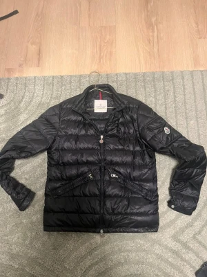Svart pufferjacka från Moncler - Snygg svart pufferjacka från Moncler med quiltad design och två dragkedjeförsedda fickor framtill. Jackan har hög krage, diskret Moncler-logga på ärmen och glansigt material som ger en exklusiv känsla. Perfekt för kalla dagar och riktigt trendig look.