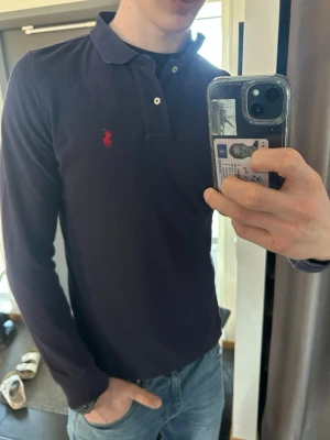 Marinblå långärmad piké från Polo Ralph Lauren - Säljer en marinblå långärmad pikétröja från Polo Ralph Lauren med klassisk krage och knappar framtill. Tröjan har det ikoniska röda Polo-logot broderat på bröstet och är gjord i mjuk bomull. Perfekt till jeans eller chinos.