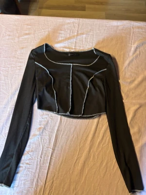 Svart croppad långärmad topp - Trendig svart croppad topp med långa ärmar och kontrastsömmar i vitt. Toppen har en ribbad struktur och markerade sömmar framtill som ger en edgy look. Perfekt för att styla med höga jeans eller kjol.
