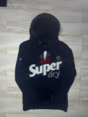 Svart Superdry hoodie med tryck - Snygg svart hoodie från Superdry med stort broderat logotyptryck i vitt, grått och rött på bröstet🤩 Hoodien är i fint skick (inga snören) och passar Xs-S🙌🏻 Hör av er vid funderingar samt snabbaffär😇