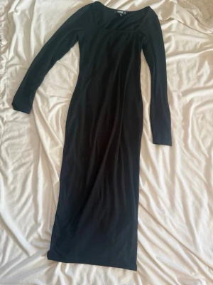 Svart långärmad långklänning - En stilren svart långklänning med långa ärmar och en något fyrkantig ringning. Klänningen har en figurnära passform och är tillverkad i ett mjukt, stretchigt material som ger en bekväm känsla.