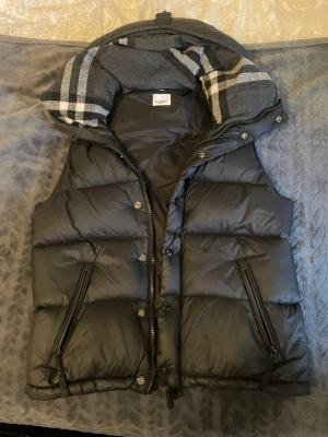 Svart dunväst från Gordon Richardson - Snygg svart dunväst  med avtagbara armar och rutigt innerfoder. Västen har två dragkedjeförsedda fickor framtill och stängs med både dragkedja och knappar. Perfekt för lager på lager och streetwear-vibe.