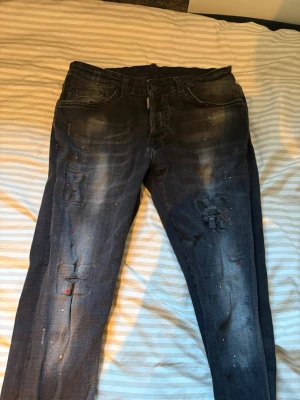 Svarta dsquared2 jeans - Säljer ett par svarta jeans med slitningar och hål på båda benen samt coola färgstänk i rött och vitt. Jeansen har klassisk smal passform. Perfekta för dig som gillar en trendig och avslappnad stil.
