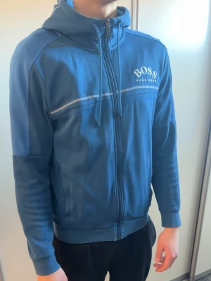 Blå hoodie från Hugo Boss - Snygg blå hoodie från Hugo Boss med dragkedja framtill och justerbar huva med snören. Perfekt för en avslappnad och stilren look.