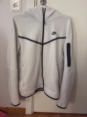 Nike - Beige/vit aktig Nike tech fleece, storlek M, säljer då den är för stor för mig. Den har en ficka på armen och två vanliga fickor, samt två innerfickor,  Nike trycket är kvar och den har svarta dragkedjor och vissa detaljer är svarta. Använd 5-8 gånger, pris kan diskuteras.💕