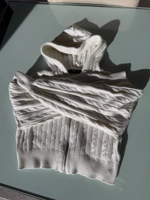 Vit stickad hoodie med dragkedja - Säljer en vit stickad hoodie med dragkedja framtill. Tröjan har ett snyggt kabelstickat mönster, huva med dragsko och ribbade muddar. Croppad modell som passar perfekt till jeans eller kjol.