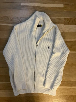 Vit stickad ziptröja Polo Ralph Lauren - Klassisk vit ribbstickad ziptröja från Polo Ralph Lauren med hög krage och broderad logga på bröstet. Tröjan har långa ärmar, dragkedja framtill och är gjord i mjukt material som passar perfekt till lager-på-lager-stil.