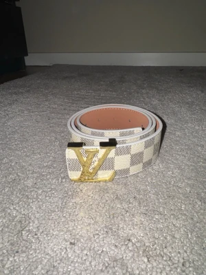 Rutmönstrat bälte med guldfärgat metallspänne - Stilrent bälte i ljust rutmönster med guldfärgat metallspänne. Klassisk design som passar både till vardag och mer uppklädda outfits. Fint skick enligt bilder.