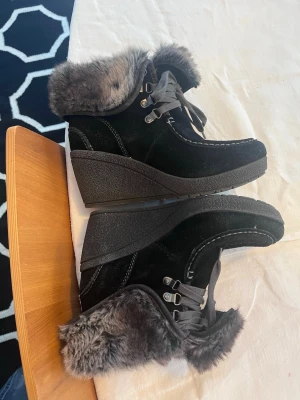 W2k stövlar med klack  - Snygga svarta stövlar från Tamaris med kilklack och grå fuskpäls runt skaftet. Snörning framtill och robust sula för extra grepp. Perfekta för kalla dagar när du vill vara både varm och trendig. Materialet är mockaimitation och detaljer i metall.