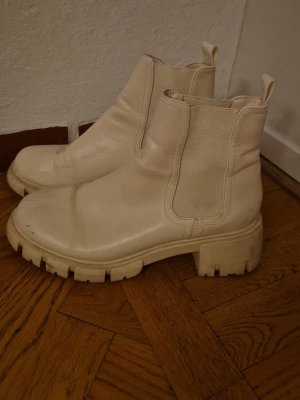 Ljusa Chunky Chelsea boots - Säljer ett par chunky Chelsea boots med bred sula och elastiska paneler på sidorna. Färg: off-white / ljust beige. Bootsens ovandel är i slätt skinnliknande materia i lite plastig känslal och de har en modern, hög siluett. Perfekta för dig som vill ha en clean och trendig look. Ena skon har en blå missfärgning, se två av bilderna, har testat gnugga lite med diskmedel.