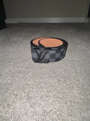 Reversibelt designerbälte – Damoflage 40mm - Exklusivt reversibelt designerbälte i stilren svart Damoflage-design med diskret pixelmönster. Ena sidan i coated canvas med mönster och andra sidan i slätt läder – perfekt för att variera din stil.  Utrustat med ett elegant svart metallspänne som ger en modern och clean look. Bredd ca 4 cm, vilket gör det till en klassisk och trendig accessoar.  • Reversibelt (2 i 1) • Svart tonad metallspänne • Premium material (canvas + läder) • Tidlös 