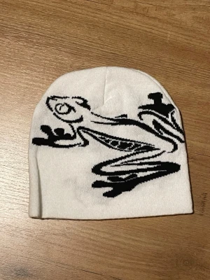 Vit mössa med svart grodmotiv - Säljer en vit mössa med ett coolt svart grodmotiv på framsidan. Mössan är mjuk och stretchig, perfekt för att hålla värmen. Priset är inte hugget i sten 