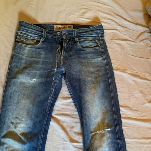 Blå Replay jeans med slitningar - Säljer ett par blå Replay jeans med snygga slitningar och tvättade detaljer. Klassisk femficksmodell med dragkedja och knapp. Jeansen har en rak passform och tydliga slitningar på låret och knät, trendigt och snyggt! Denna modell säljs inte längre (retro).