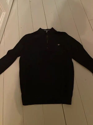 Svart stickad halvzip från Gant - Mörk mörk blå stickad tröja från Gant med halv dragkedja och broderad logga på bröstet. Tröjan har lång ärm och ribbade muddar vid ärmslut och nederkant. Perfekt för dig som gillar en stilren och klassisk look.