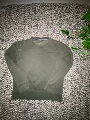 Olivgrön sweatshirt från Sail Racing - Olivgrön crewneck sweatshirt från Sail Racing med diskret logga på bröstet. Tröjan har rund halsringning, ribbade muddar och mjukt fleecefoder på insidan. Perfekt för dig som gillar stilrena och bekväma plagg.