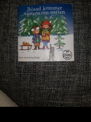 Ibland kommer vintern om natten - En liten fyrkantig Pixibok med titeln 'Ibland kommer vintern om natten' av Karin Stjernholm Ræder. Omslaget är blått och vitt med illustration av två barn i vinterkläder, en katt och en pulka framför snötäckta granar. Perfekt för dig som gillar mysiga vinterberättelser.