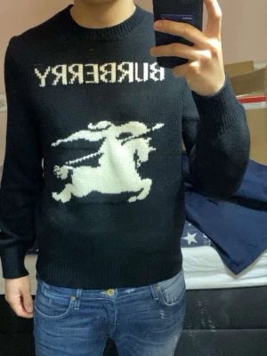 Svart stickad tröja från Burberry - Svart stickad tröja från Burberry med vit logga och motiv av riddare på bröstet. Tröjan har rund halsringning och ribbade muddar vid ärmslut och nederkant. Perfekt för dig som gillar klassisk design med en twist.