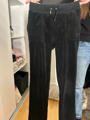 Svarta velourbyxor från Juicy Couture - Säljer ett par klassiska svarta mjukisbyxor från Juicy Couture i mjuk velour. Byxorna har dragsko i midjan, fickor fram och bak med knappar samt diskret broderad logga fram. Perfekta för en chill och trendig look.