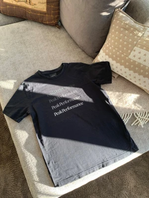 Svart Peak Performance t-shirt - Säljer en svart t-shirt från Peak Performance med flera logotryck på framsidan. Klassisk rund hals och korta ärmar, perfekt för dig som gillar stilrena plagg med tydlig branding. Materialet är mjuk bomull som känns skönt mot huden.