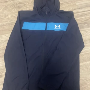 Blå vindjacka från Under Armour - S Snygg marinblå vindjacka från Under Armour med huva och dragkedja. Jackan har en bred blå panel över bröstet med logga, meshfoder på insidan och långa ärmar. Perfekt för dig som gillar sportig stil och vill ha något lätt och funktionellt.