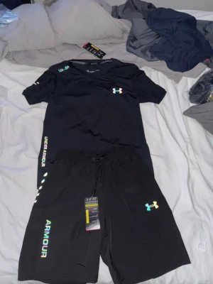 Svart träningsset från Under Armour - Snyggt svart träningsset från Under Armour med t-shirt och shorts. Setet har färgglada loggor och textdetaljer på både tröja och byxa. Materialet är lätt och andas, perfekt för gymmet eller löprundan. Elastisk midja med dragsko på shortsen för skön passform.