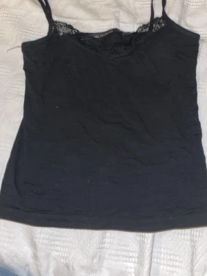 Svart linnetopp med spetsdetalj - Snygg svart linnetopp från Label Collection med tunna axelband och spetsdetalj vid urringningen. Perfekt att styla lager på lager eller bära som den är. Enkel och stilren design som passar till mycket.
