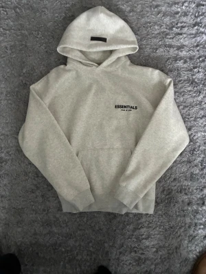 Grå Essentials hoodie - Storlek S, äkta, ny med etiketter Snygg ljusgrå hoodie från Essentials med svart logotyp på bröstet. Tillverkad i mjukt och bekvämt material, perfekt för en avslappnad stil. Passar dig som gillar streetwear och enkel design.