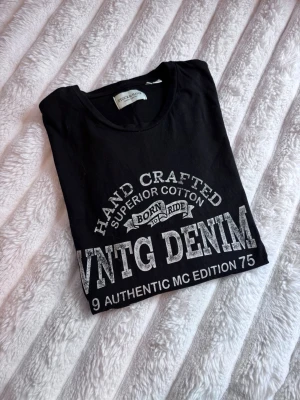 Svart t-shirt från Stock & Hank, koszulka - Snygg svart t-shirt från Stock & Hank med stort vitt tryck på bröstet där det står 'VNTG DENIM' och andra coola detaljer. Tillverkad i mjuk bomull och har klassisk rund hals och korta ärmar.  Längd på T-shirt: 65cm Axelbredd: 44cm