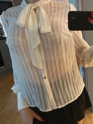 Vit transparent blus med knytband - Stilren vit blus i transparent, lätt material med vertikala ränder och knytband i halsen. Blusen har knappar framtill och långa ärmar med breda manschetter. Perfekt för en chic och trendig look.