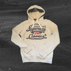 Fet Superdry Hoodie  - Ljusgrå hoodie från Superdry med trycket 'Superdry JPN Classics' i marinblått och rött framtill. Hoodien har huva med vita dragsnören och en klassisk känguruficka. Tillverkad i mjukt bomullsmaterial, perfekt för en avslappnad streetwear-look.