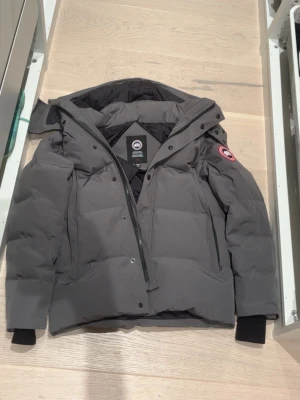 Grå dunjacka från Canada Goose - Snygg grå dunjacka från Canada Goose med avtagbar huva och klassisk logga på ärmen. Jackan har dragkedja och knappar framtill, samt två sidofickor. Perfekt för kalla vinterdagar tack vare det tjocka dunfodret och vindtäta materialet.