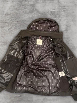 Svart pufferjacka från Moncler - Säljer en snygg svart pufferjacka från Moncler i mycket bra skick. Jackan har en quiltad design med dragkedja och huva, perfekt för kyliga dagar. Insidan har ett stilrent mönster och det finns praktiska fickor med dragkedjor. Passar perfekt för både höst och vinter!